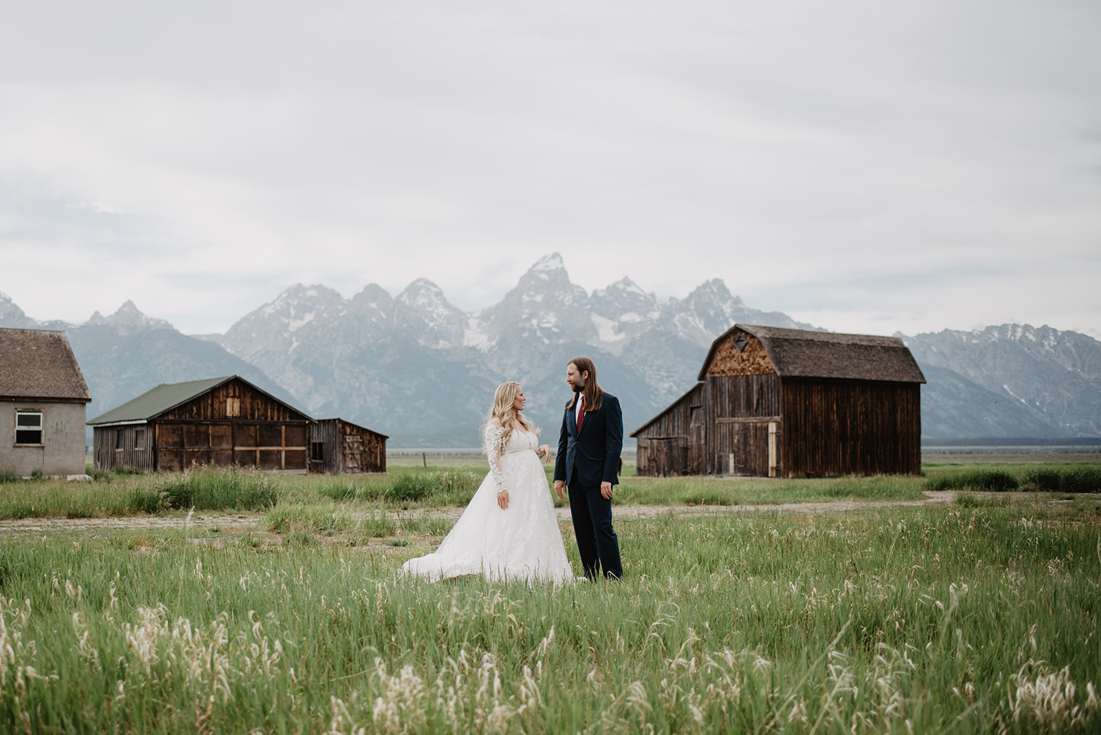 bride in a large flowy dress with her groom in a black suit stand together in a pasture infron of a barn and hold hands while looking at each other