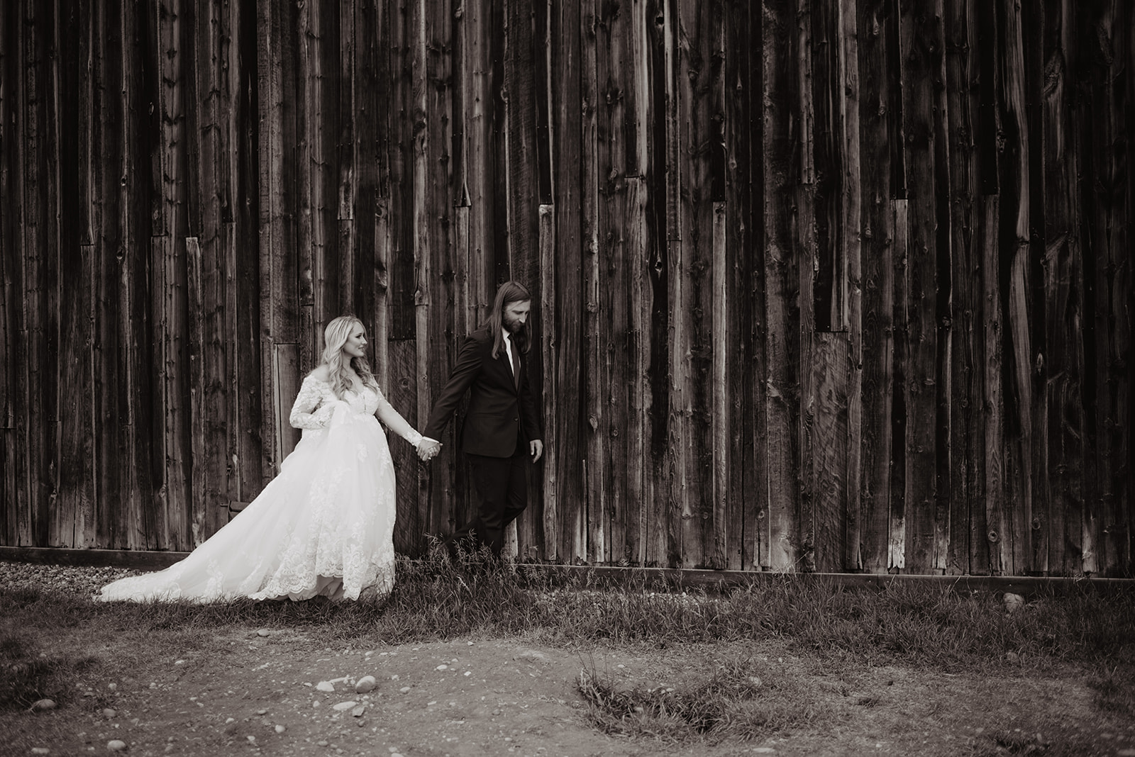 black and white photo of bride holding grooms hands as he leads her on a dirt path next to a barn in Jackson Hole on their wedding day