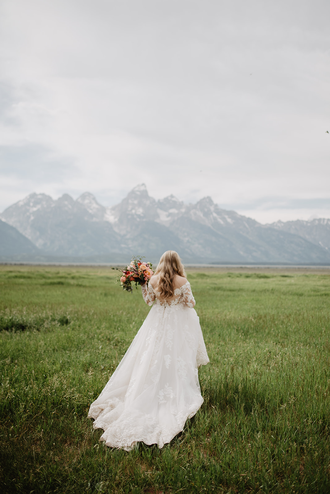 blonde bride with soft curls walks through a green field towards the Tetons while holding her whimsical dress in one hand and her bridal bouquet in another during her Jackson Hole wedding at Diamond Cross Ranch
