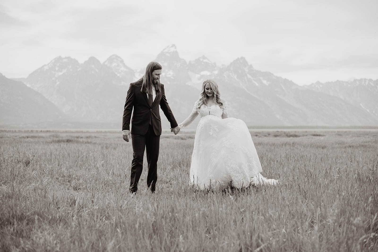 black and white photo of bride holding grooms hand and smiling as she twirls in her wedding dress in a field with the Grand Tetons behind her