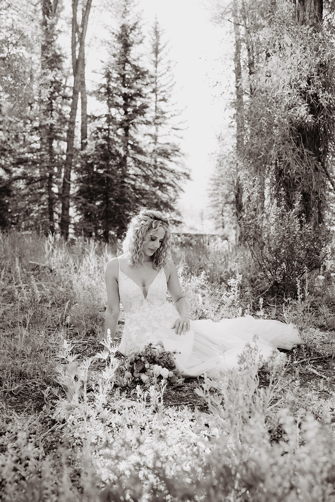 black and white photo of bride with light curly hair sitting in a meadow in the woods with florals surrounding her and tall trees towering over her
