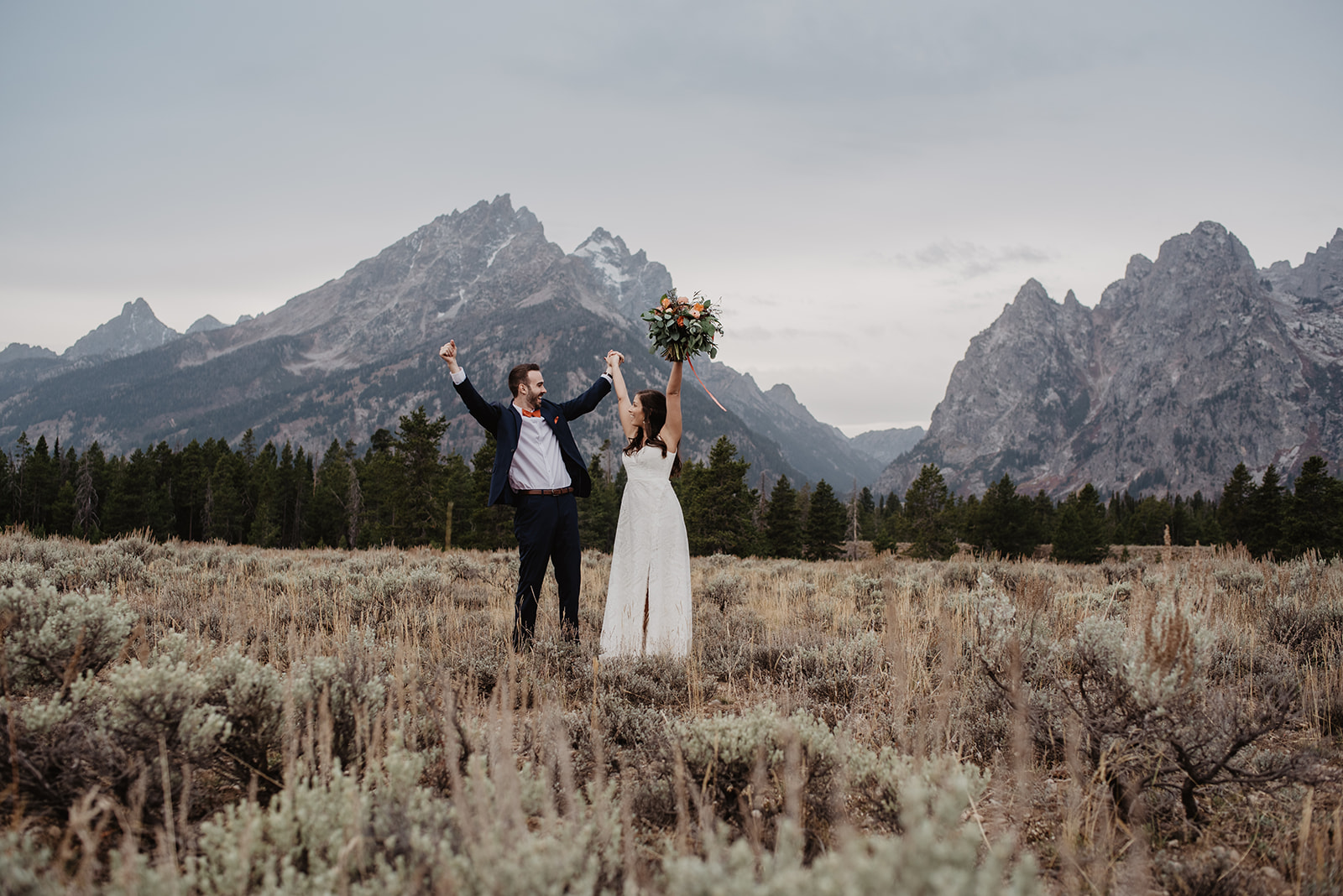 Jackson Hole wedding day with bride and groom standing in front of the Grand Tetons with their hands in the air as they are excited to be married