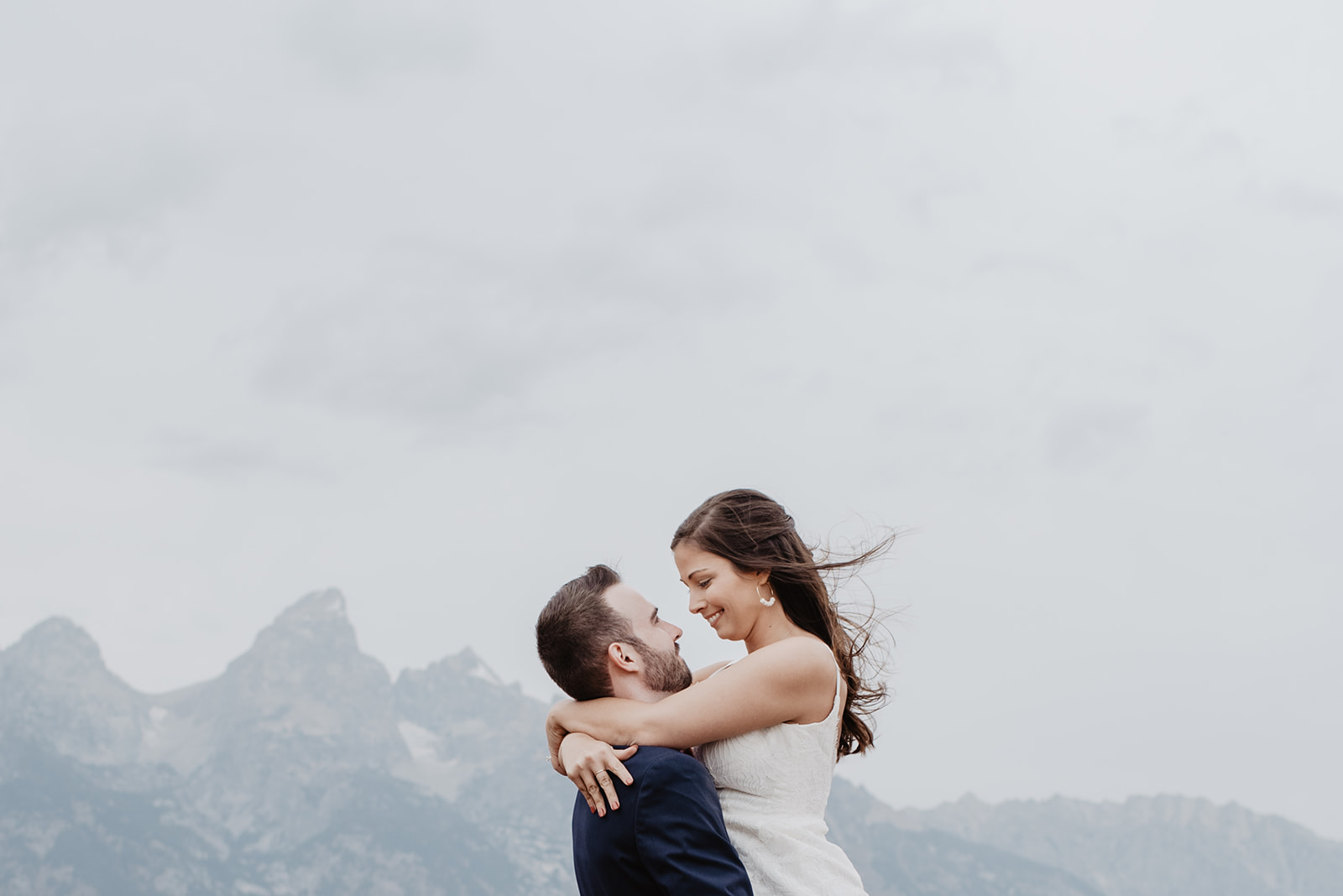 bride in a simple and elegant white gown is being picked up by her groom with the wind blowing her hair and the Tetons in the background of their Jackson Hole wedding venue
