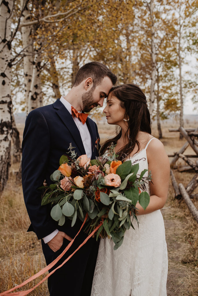 bride and groom standing close to one another with their foreheads touching in a fall forest with bride holding a warm toned wedding floral bouquet in Jackson Hole, best venues for weddings in Jackson Hole