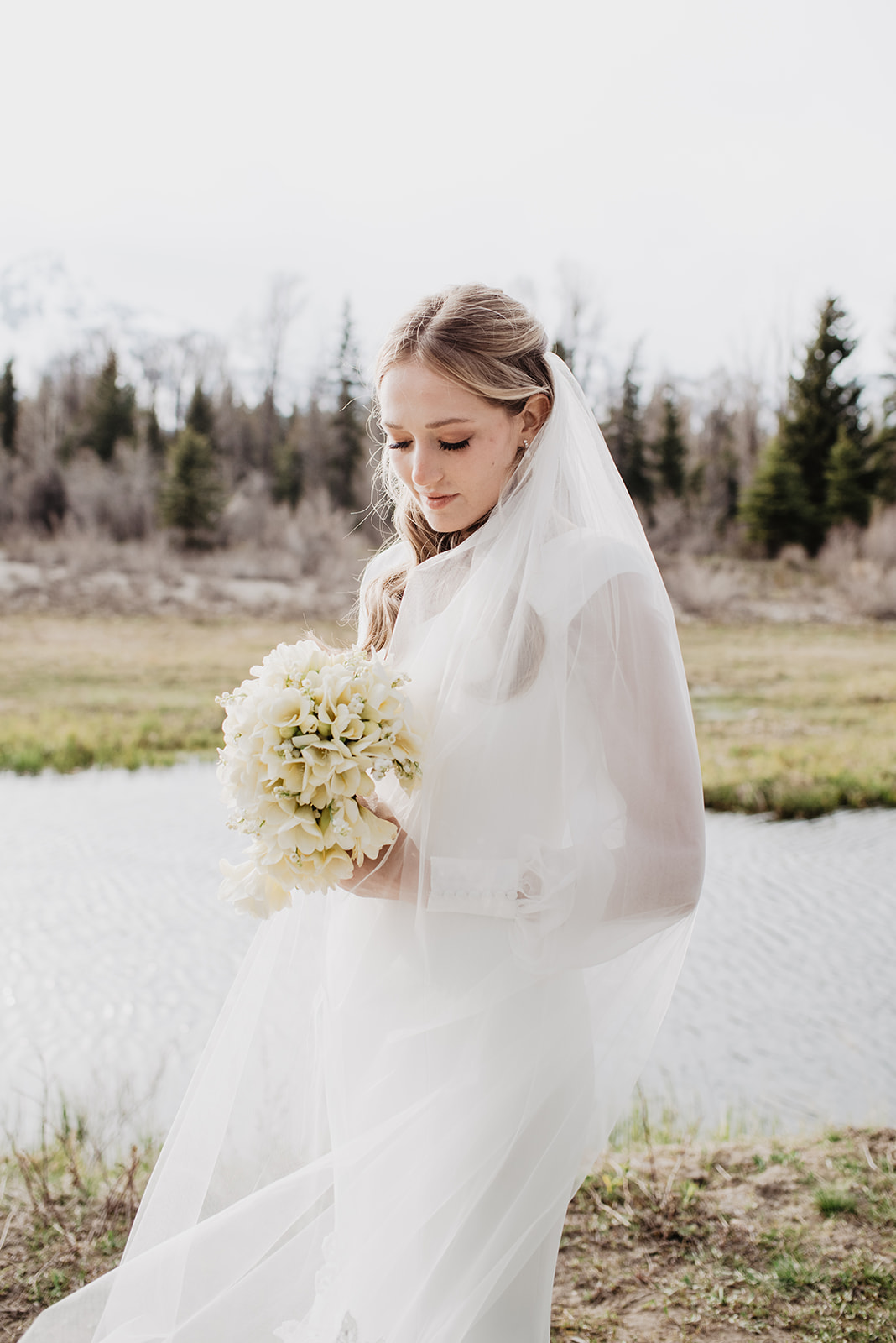 beautiful blonde bride wearing a stunning classic victorian wedding gown stands with her white lily wedding bouqut in the Tetons for her bridals