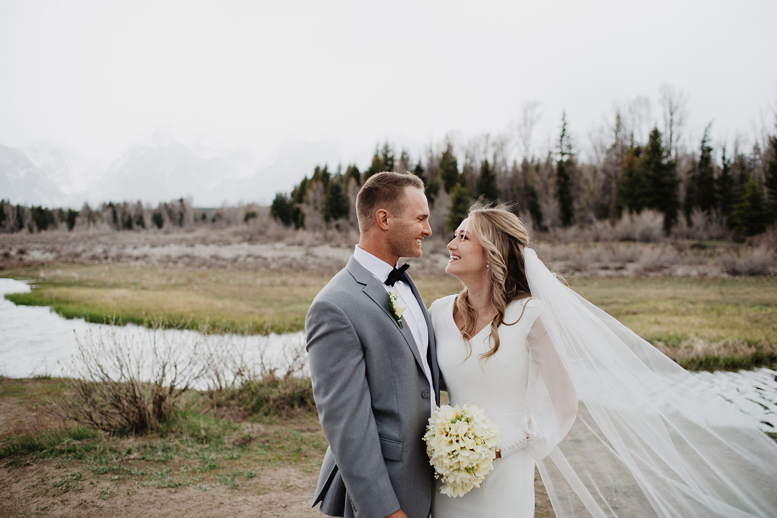 bride and groom on their foggy Jackson hole wedding day standing close together and smile while gazing at each other with the Tetons in the background through the thick fog