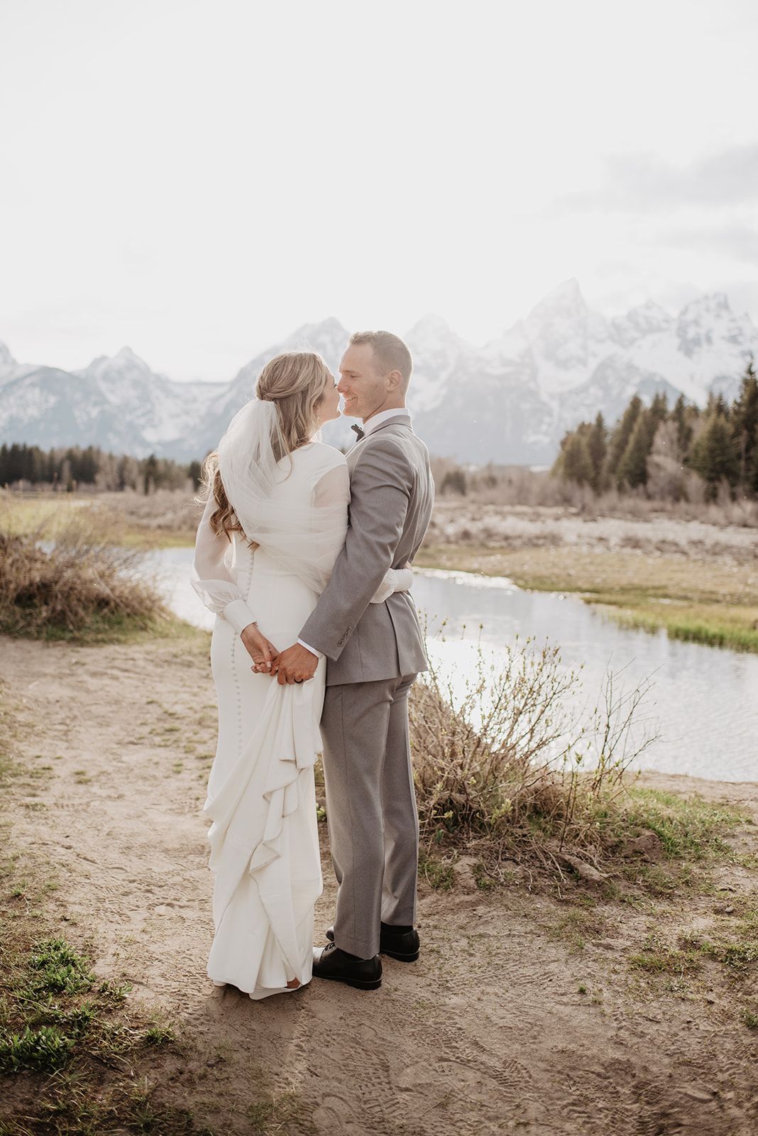 bride and groom hold hands behind their backs and lean into one another to kiss while standing in front of the Grand Tetons