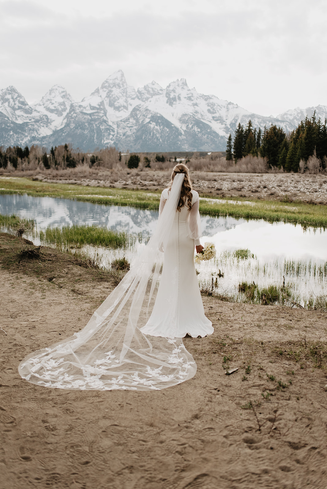 Jackson Hole bride walking towards a river in the Tetons with her long lace veil trailing behind her and the Tetons in the back ground