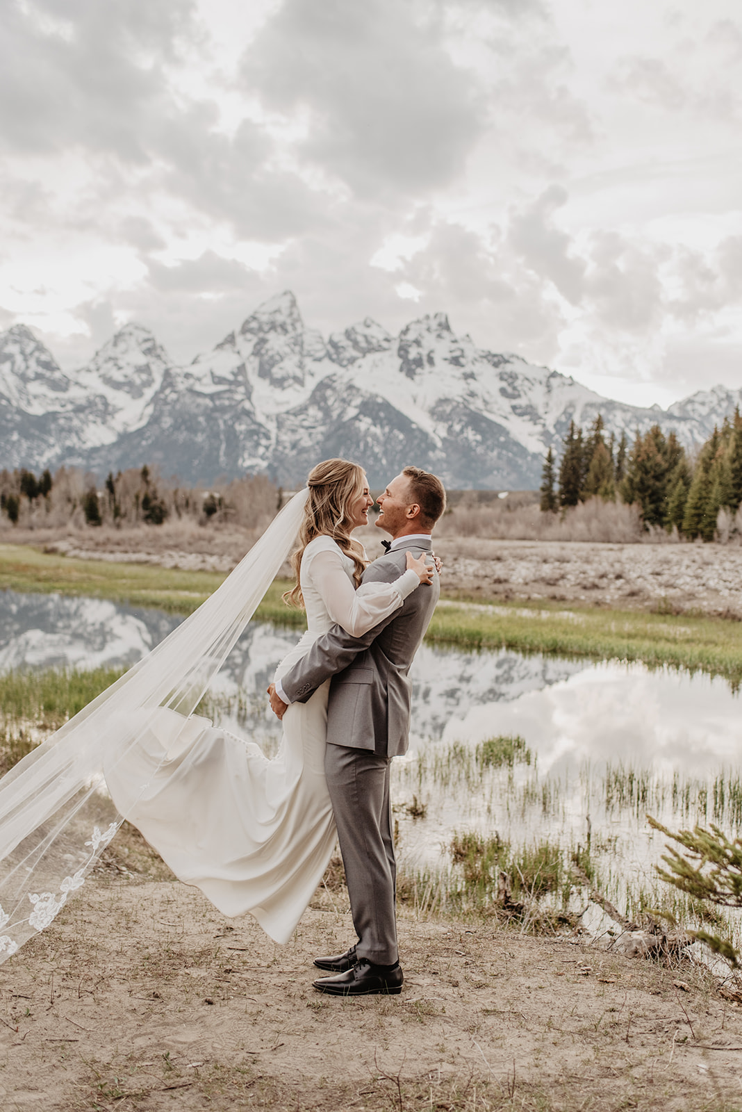 Man lifes up bride from her legs as she smiles and her wedding veil flows behind her in front of the Tetons for their Jackson Hole wedding
