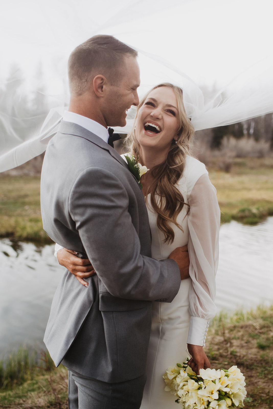dreamy wedding day at Jackson Lake Lodge with bride laughing and groom smiling as he holds her waist in fron tof a river in JAckson Hole with the Tetons in the background