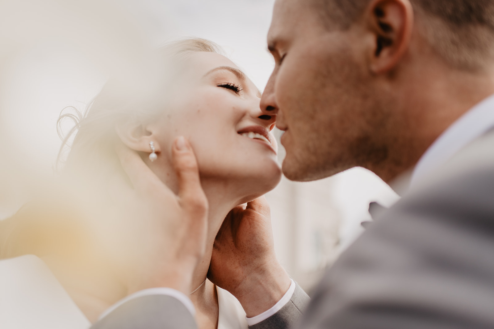 close up image of bride smiling as her groom holds her jaw lightly and leans into a kiss with glimer of sunlight coming through behind them
