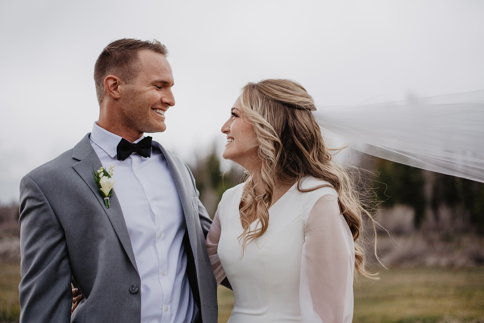 bride and groom on a foggy day in the Tetons celebrating their wedding while wearing a traditional long sleeve gown and a blue suit with a bow tie holding hands and smiling at one another