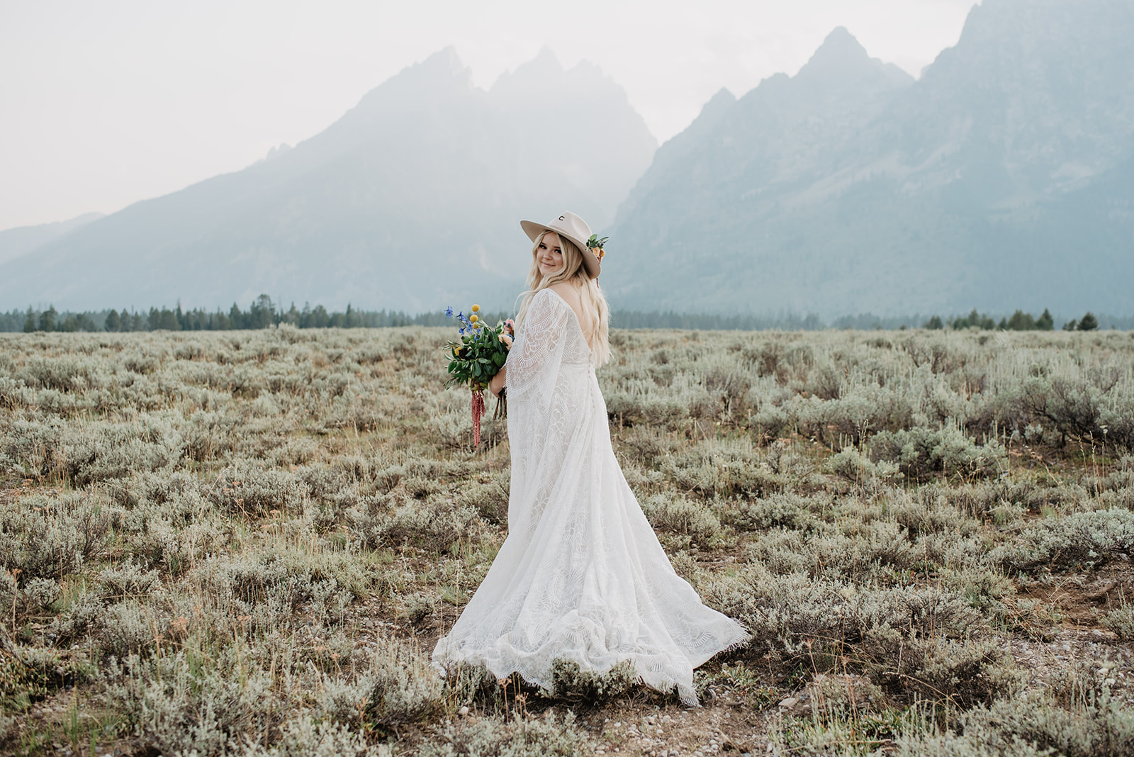 Bride posing in front of the Grand Tetons on her Jackson Hole wedding day with her back to the camera as she looks over her shoulder to the camera and holds her bridal bouquet to her side, Jackson Hole wedding venues