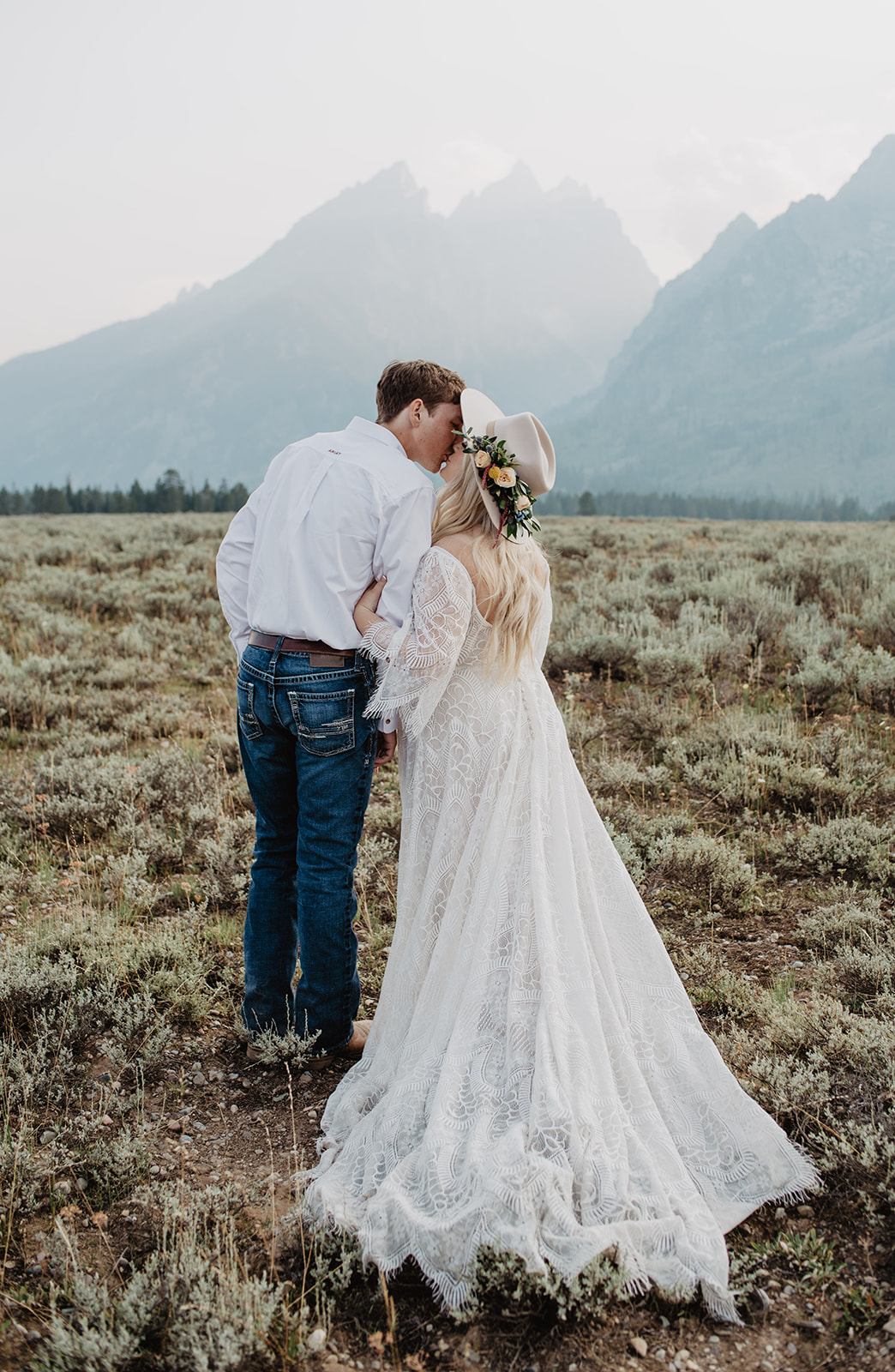 bride and groom kissing in a field in the Tetons for their bridal photos in the fall. bride wearing a bohemian lace dress with a tan hat and flowers, groom wearing jeans and a white shirt
