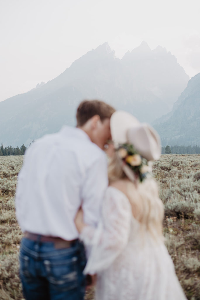 adventure elopement with young couple at the teatons with bride wearing a trendy wide brimmed hat with florals on it leaning into kiss her groom as the focus of teh picture is on the mountains in front of them