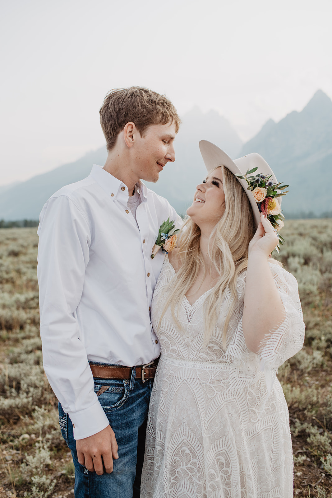 bride and groom stand together in a field near the Grand Tetons looking at one another and smiling, the bride is wearing a trending hat with florals on the side of it.