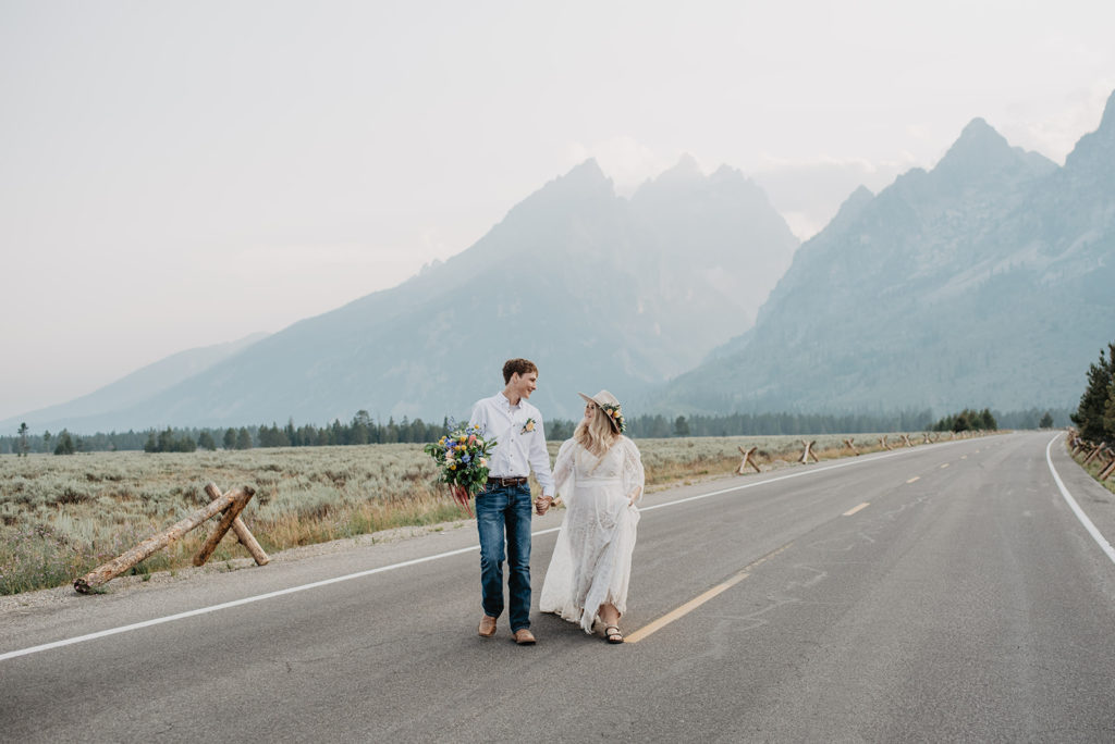 bride and groom walk down a road together while holding hands and looking at each other with the Tetons on the background
