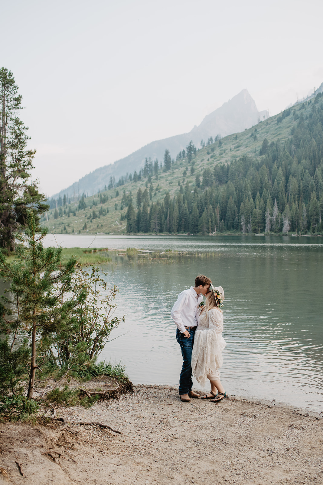 Grand Teton NAtional Park elopement with bride in a lace gown and tan hat kissing her groom in front of a lake with a mountain view