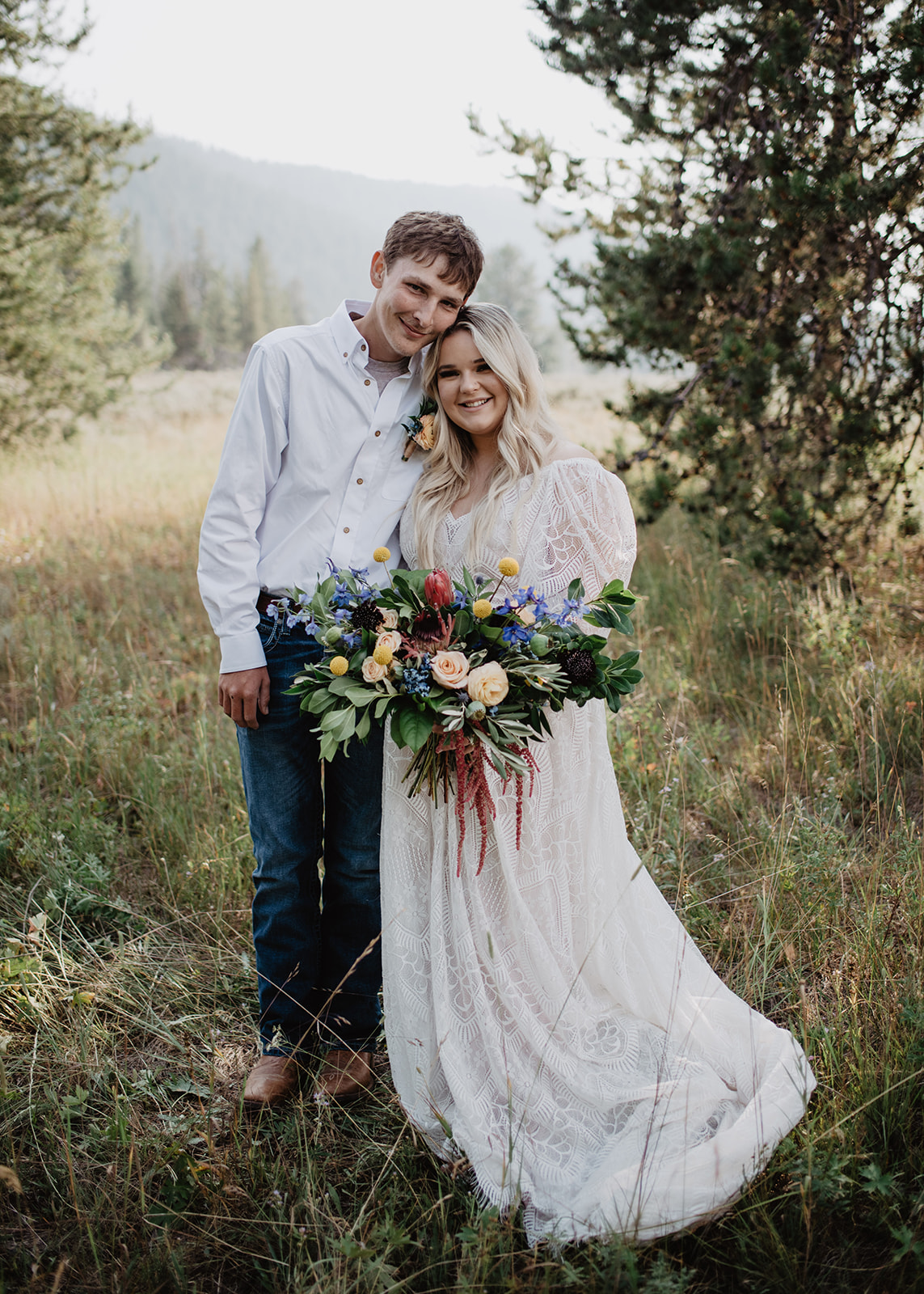 bride and groom standing next to each other in the woods at the Grand Tetons with the bride holing her wedding day bouquet as they lean into each other smile