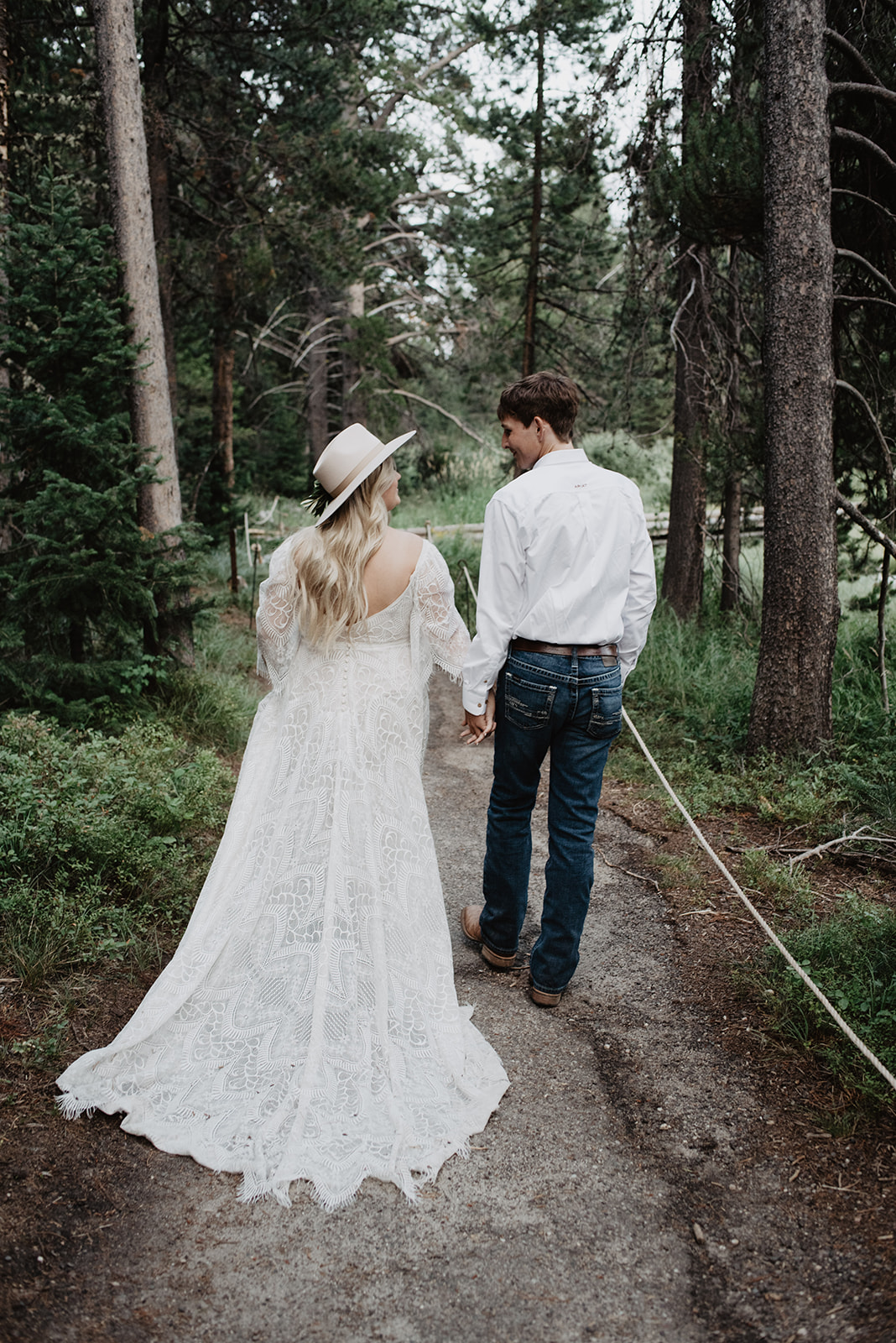 young couple walk away from the camer into the woods on a dirt path while holding hands and looking at each other with tall trees surrounding them in the Grand Tetons