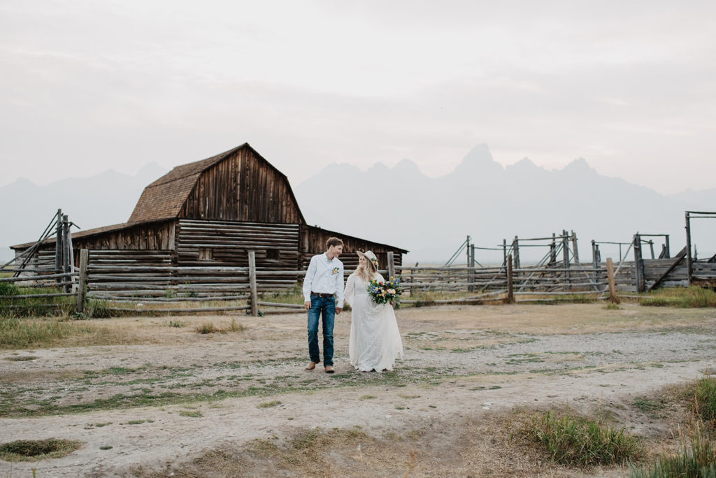 Jackson Hole photographer captures wyoming elopement with man and woman holding hands and walking across a dirt path in front of an old historic barn 