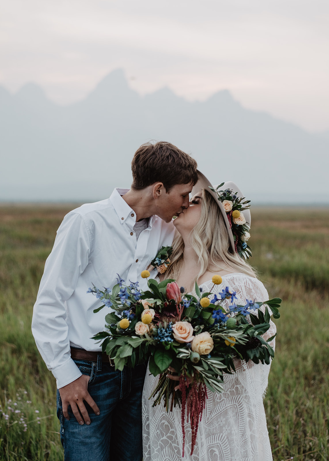bride and groom kissing in a field in Grand Tetons with a hazed sky and a light sunset behind them and the silhouette of the Tetons.