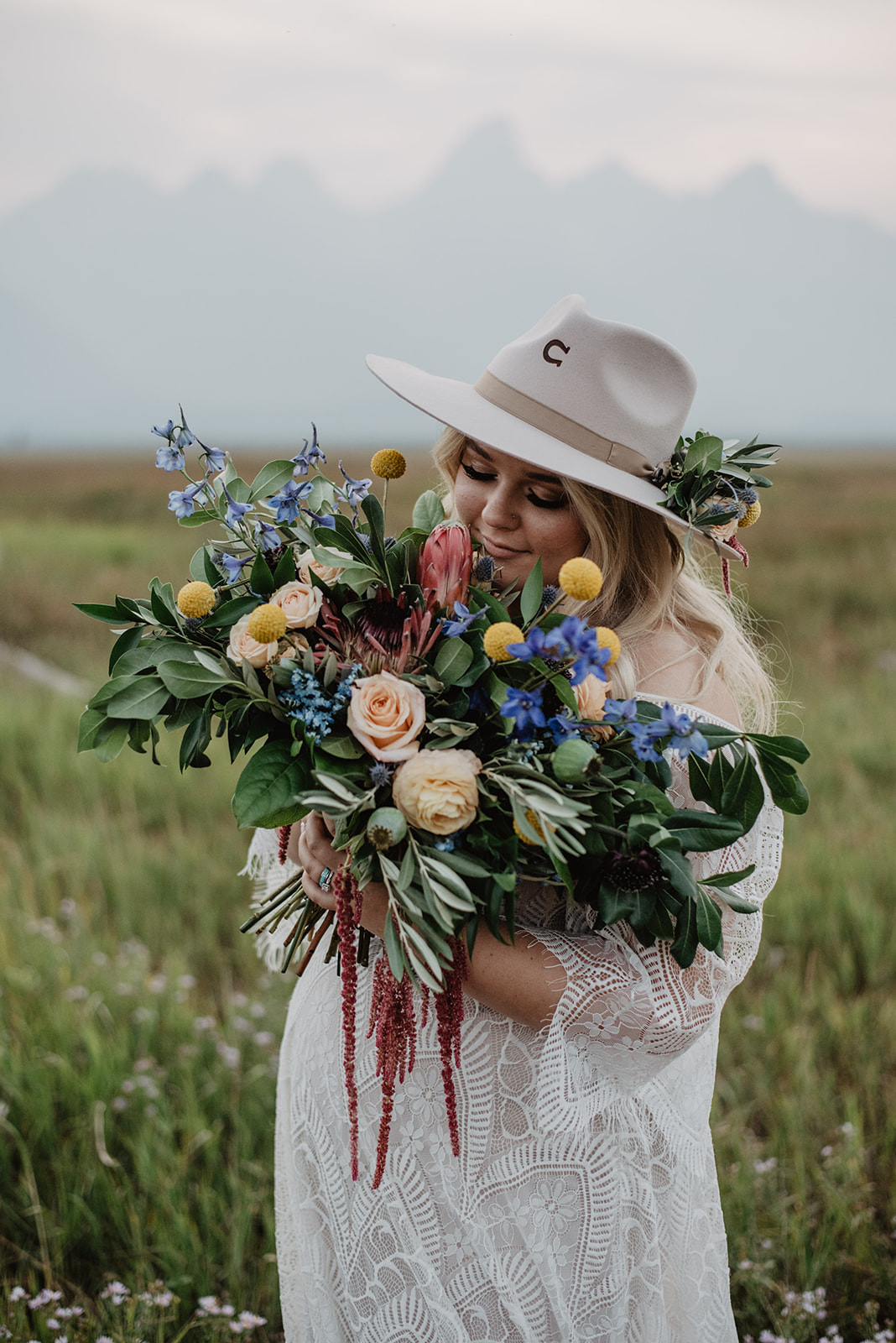 brides holding her wedding flowers up to her face and smelling them with the Grand Tetons behind her as she wears a boho lace wedding dress and a tan hat