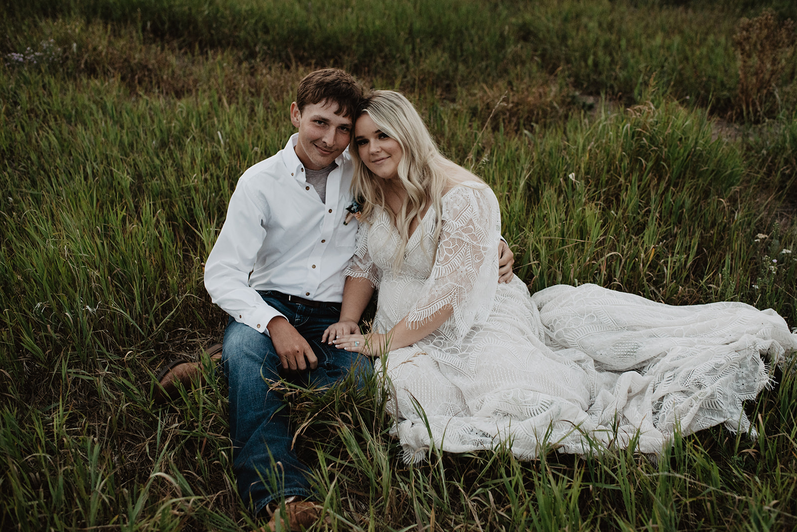 wedding photo in the Grand Tetons with man and woman sitting in a grass frield together holding hands and smiling with their heads tilted together