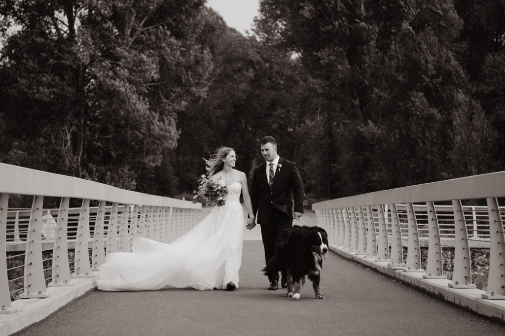 Jackson Hole photographers capture bride and groom walking their dog on a bridge to a lake in the tetons with the woods behind them captured by jackson hole wedding photographer