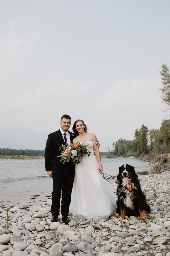 Jackson Hole photographers capture bride and groom standing on a river rock bed with their dog in the Tetons for their elopement