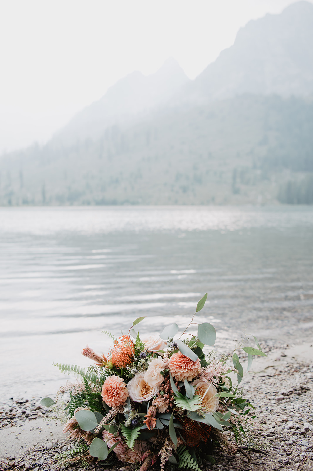 beautiful coral wedding bouquet sitting next to String Lake in the Tetons with a mist over the water behind the florals