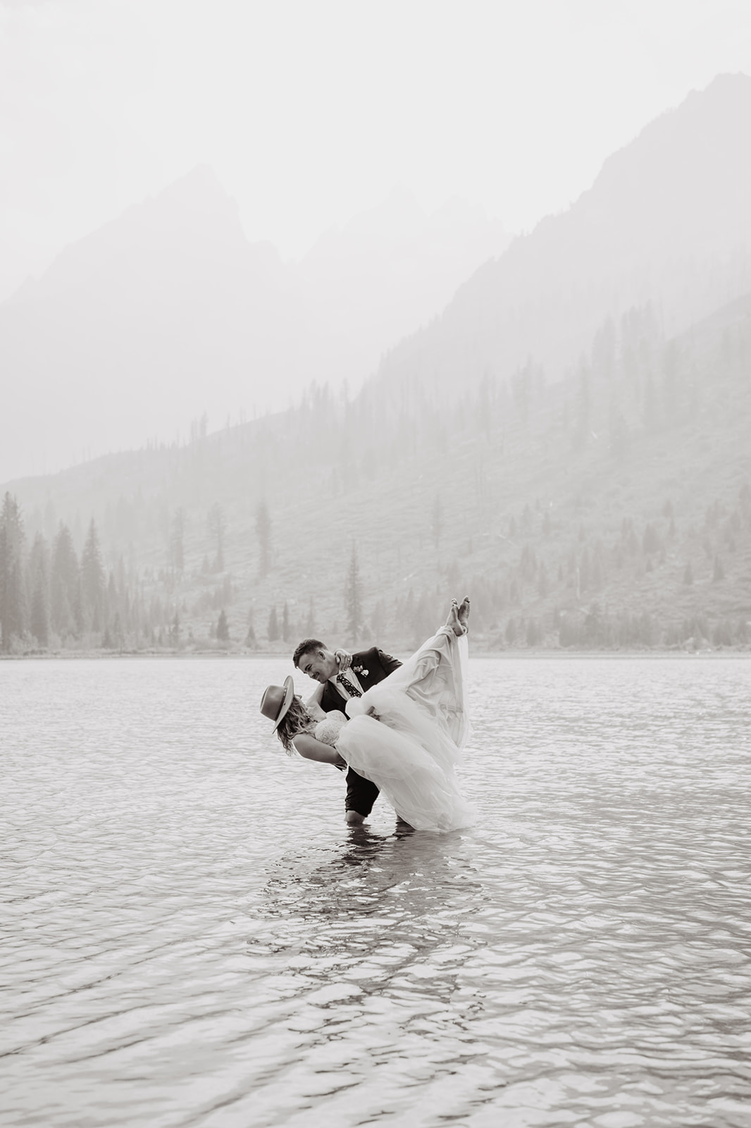 black and white image of husband and wife on their wedding day the Grand Tetons, the groom carrying his wife in teh water and dippin her back as if they are dancing together in the lake