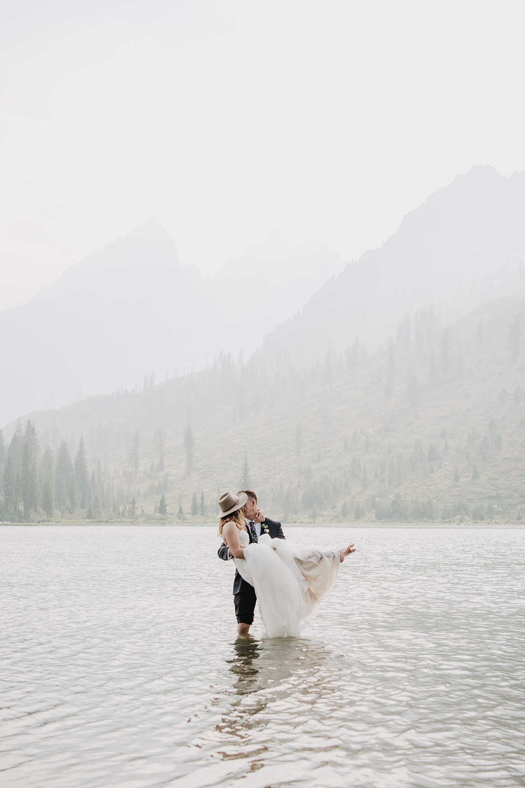 adventure elopement in Jackson Hole with bride and groom in the water at String Lake with the groom carrying the bride and her dress flowing from the wind into the water