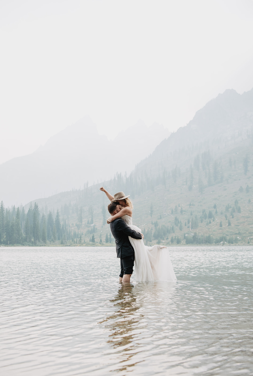 bride and groom stannding in String Lake in the Tetons together as the grom who is in a black suit lifts his bride up in the air and the bride whois in a lace and tulle wedding dress kicks up her foot behind her and throws her fist into the air in victory