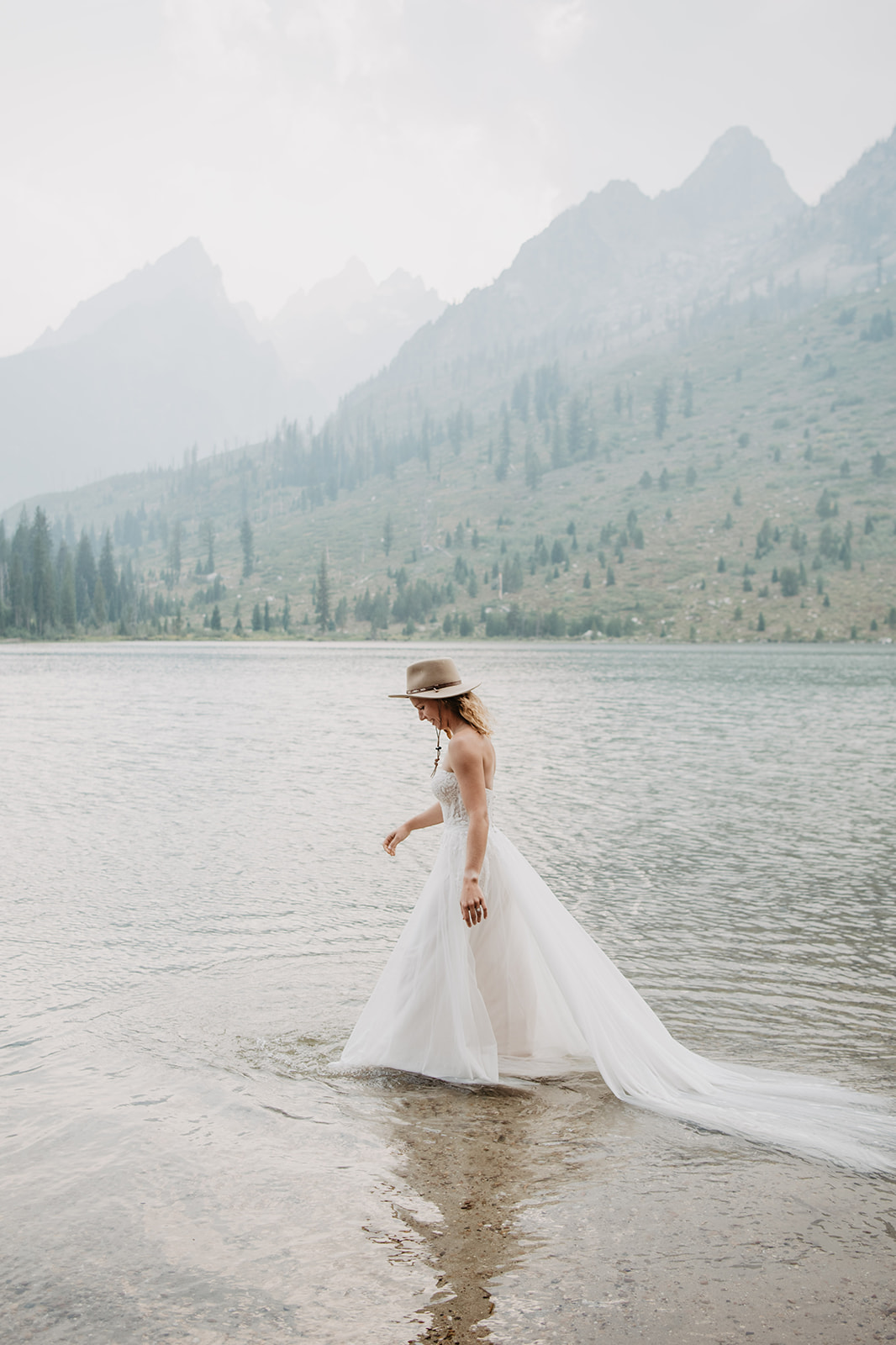 String Lake bridals in Jackson Hole with adventurous bride walking through the water with her wedding dress and a tan hat on
