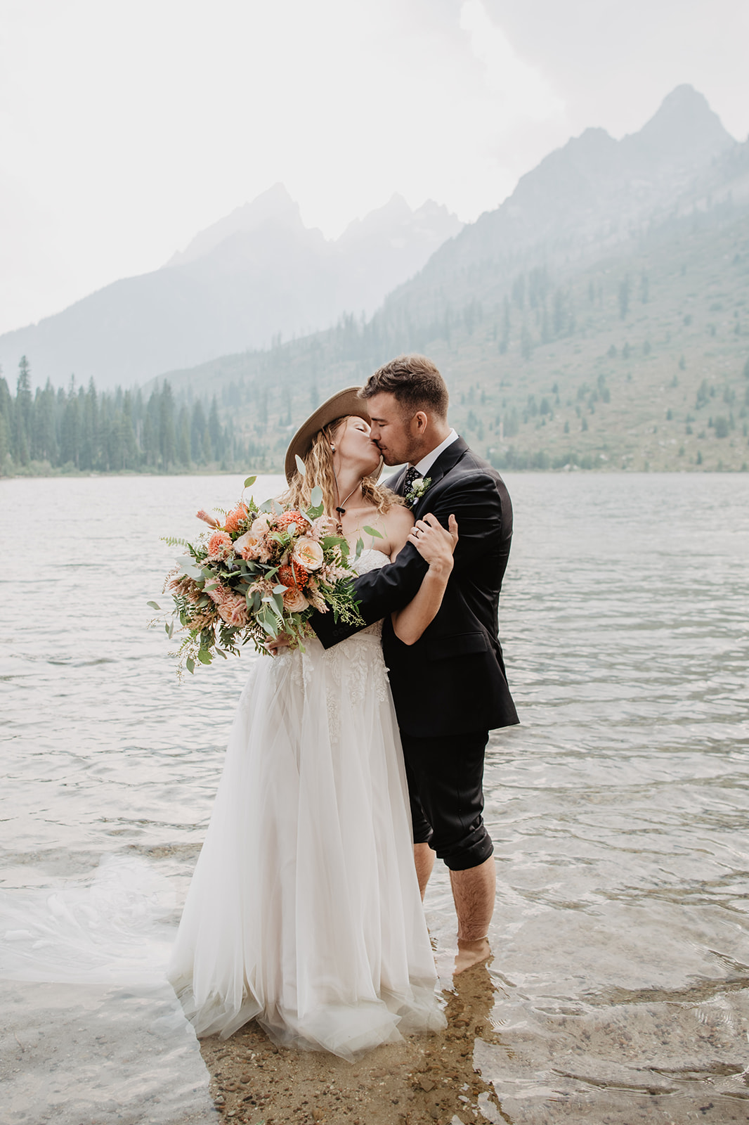 Jackson Hole elopement with bride and groom standing the lake and kissing while the bride wears a tan hat and holds her wedding florals as her groom kisses her passionately
