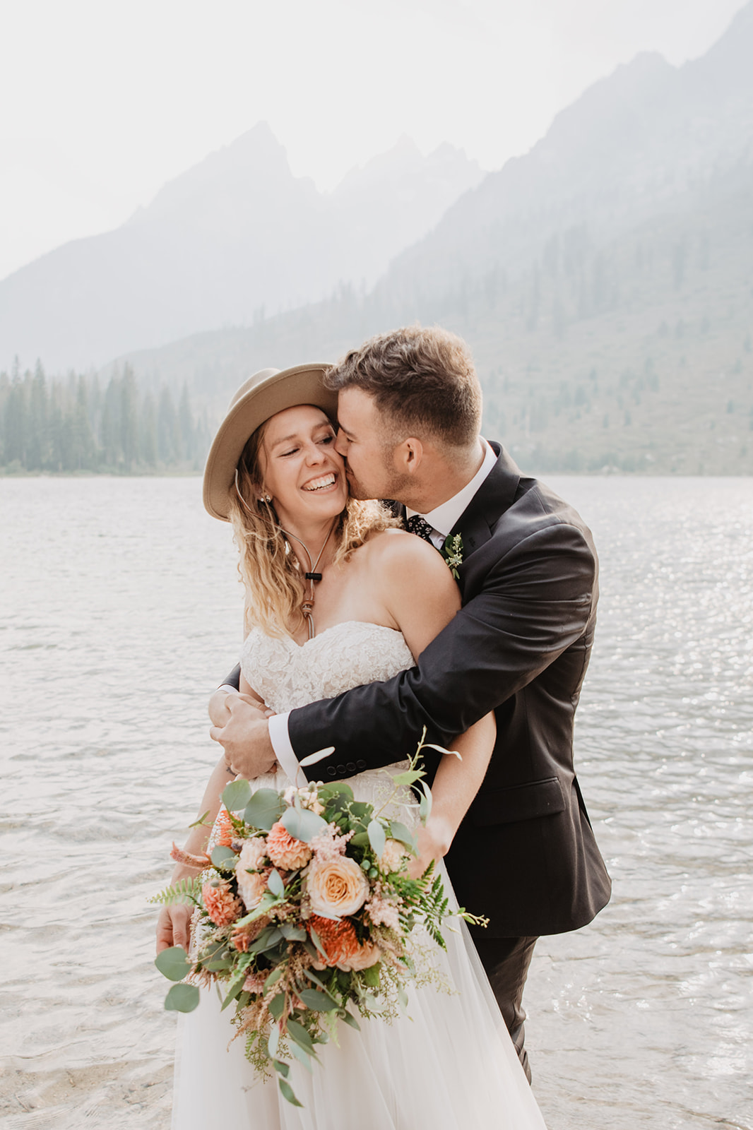 Jackson Hole wedding photographer capture bride and groom in a lake embracing each other while the groom kisses his bride as she laughs while holding her florals 