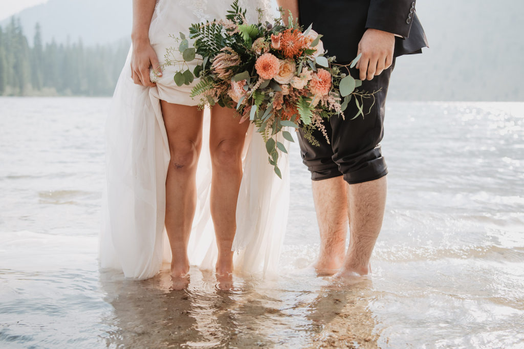 bride and groom walking through the water at a lake in Jackson Hole with the Tetons in the distance as the bride holds up her wedding dress captured by Jackson Hole photographers