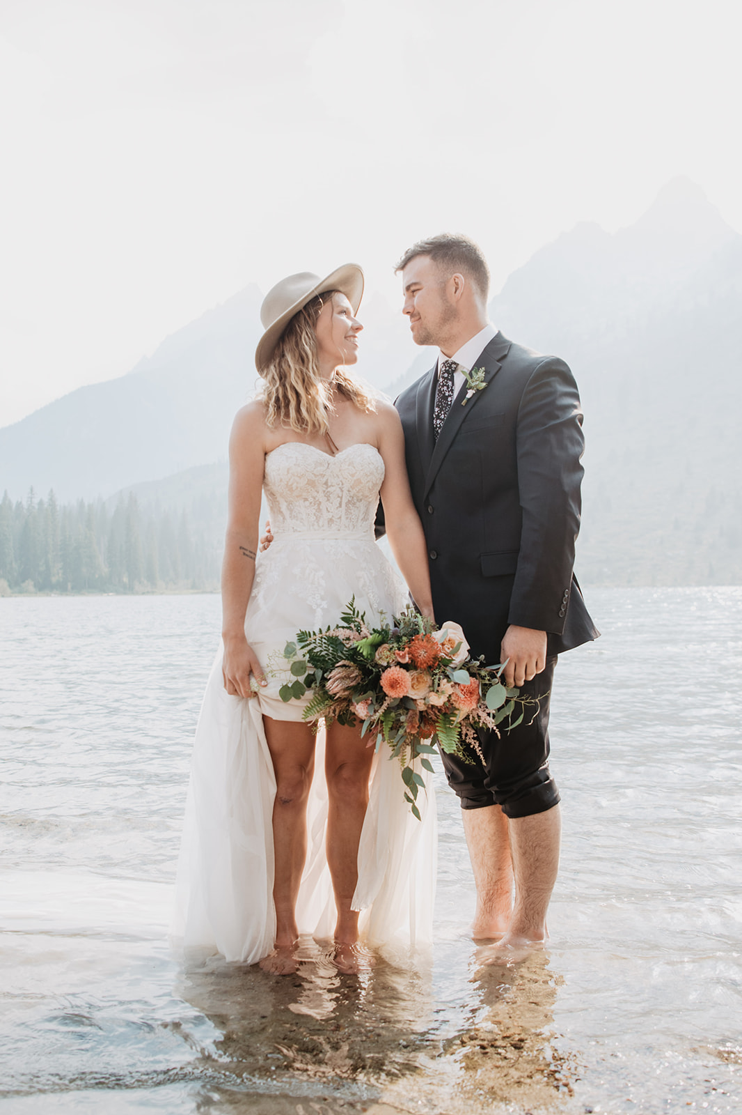 bride in a tulle and lace strapless a-line wedding dress holds up her skirt, as well as her coral wedding bouquet while wearing a hat as she looks at her groom as his pants are rolled up and they stand in Jackson Hole's String Lake together