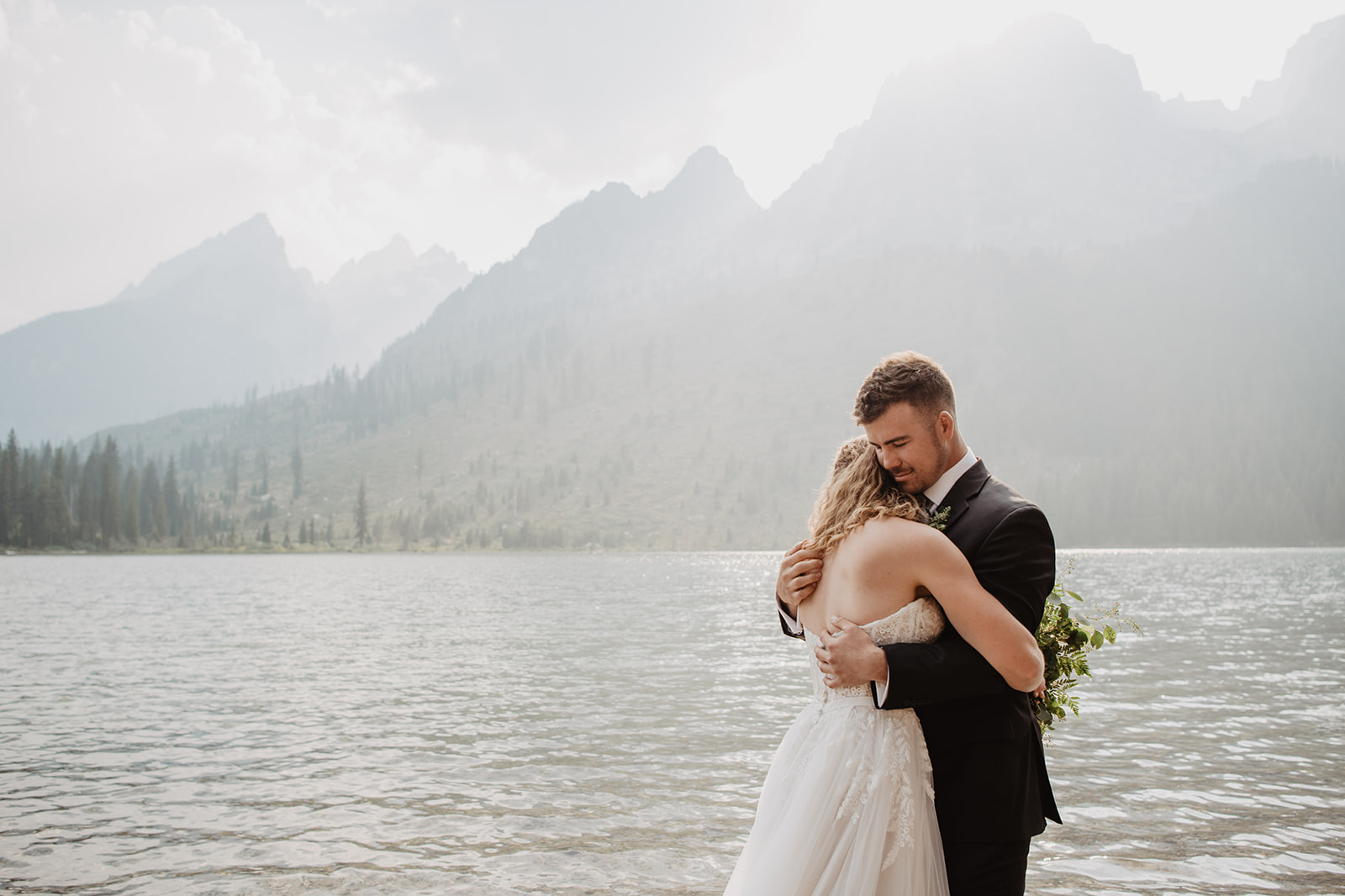 groom in a black suit holds his bride as they both stand in String Lake in Jackson Hole with the Tetons right behind them as the sun glows through the smoke that is in the air