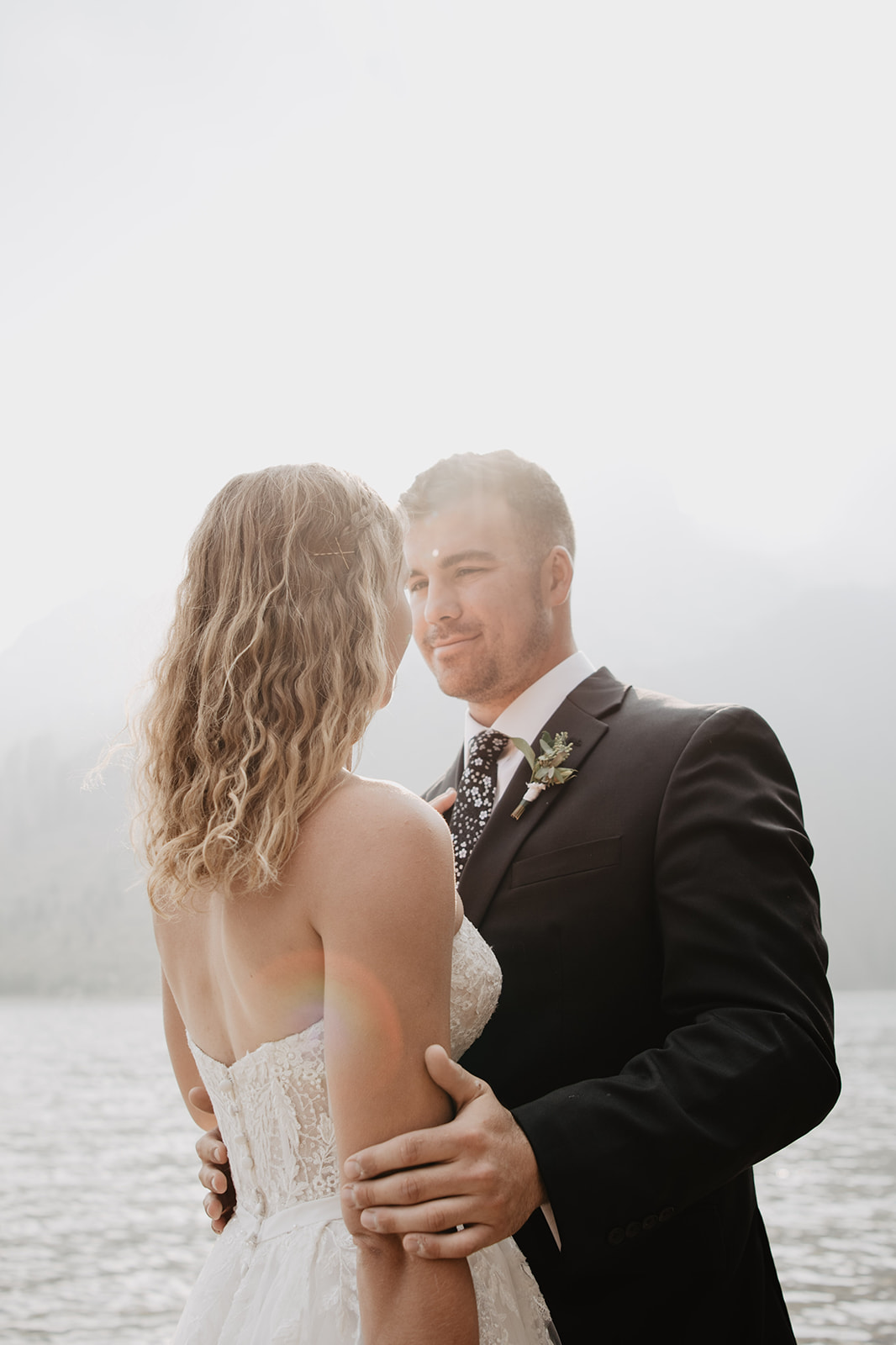 groom holding his bride as the sun shines over head with the water in the background and mountains behind them