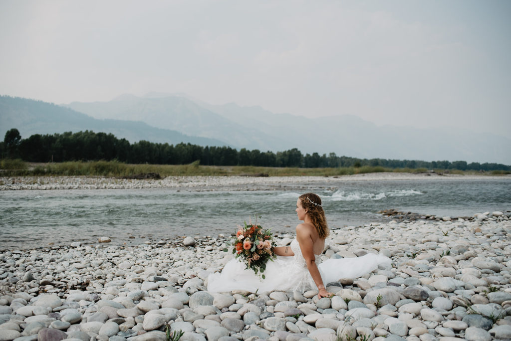 bride sitting on a rock beach in jackson hole for bridal photos looking out to the water while holding her bouquet 