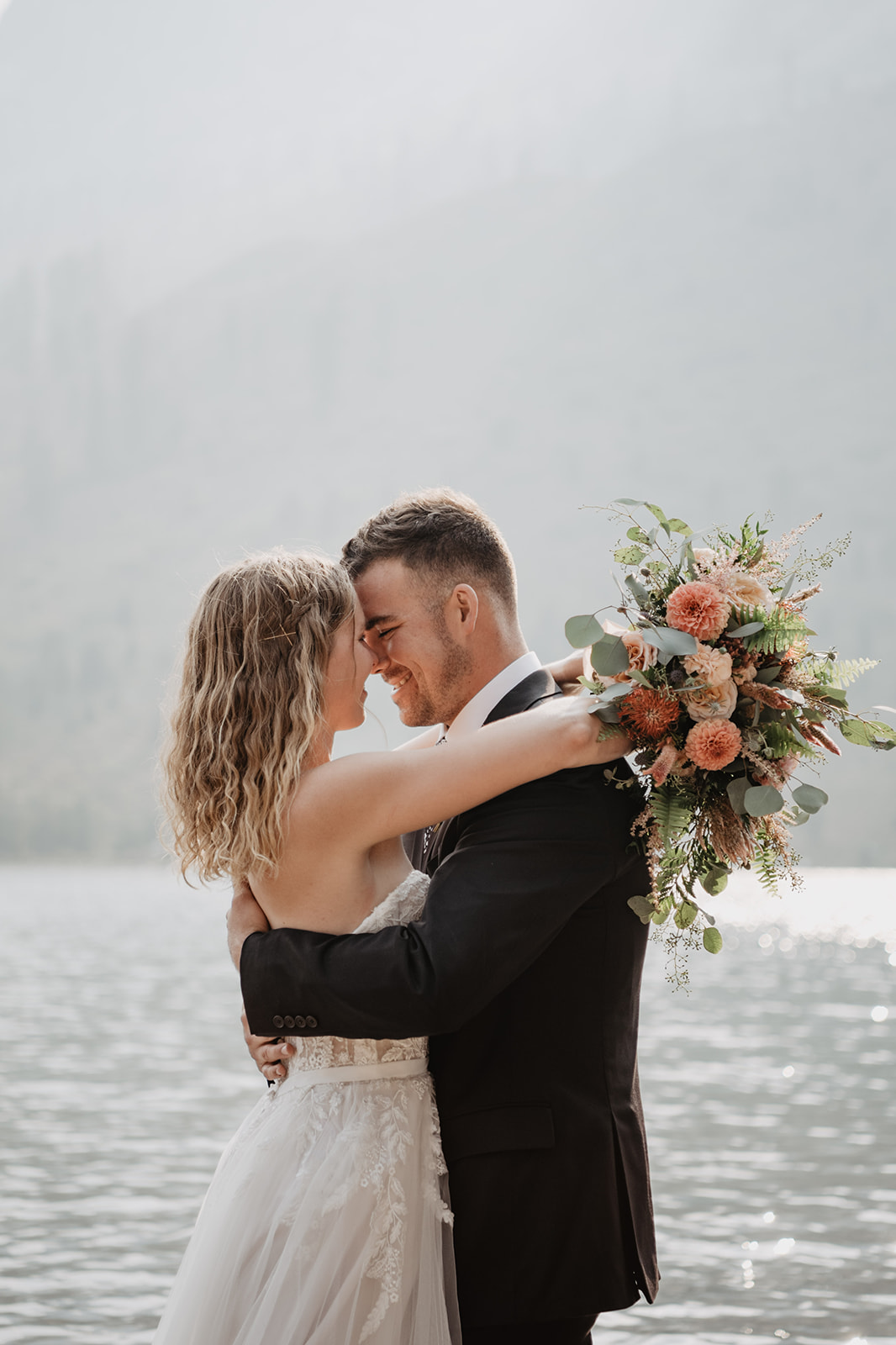 bride and groomin the Grand Tetons planning to elope as they stand at a lake embracing each toher and looking deeply into one another's eyes while the bride holds her pink and cream wedding bouquet over the groom shoudlers