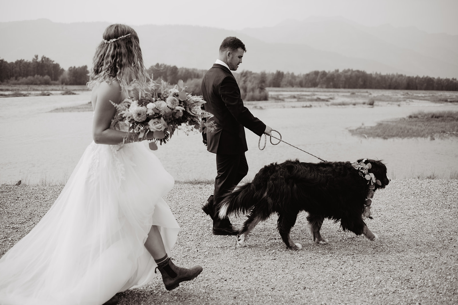 black and white photo of bring and groom walking their dog in Jackson Hole along a trail next to a lake with bride holding her wedding bouquet and groom holding the dog's leash. Bride wearing hiking boots and her tulle and lace wedding gown