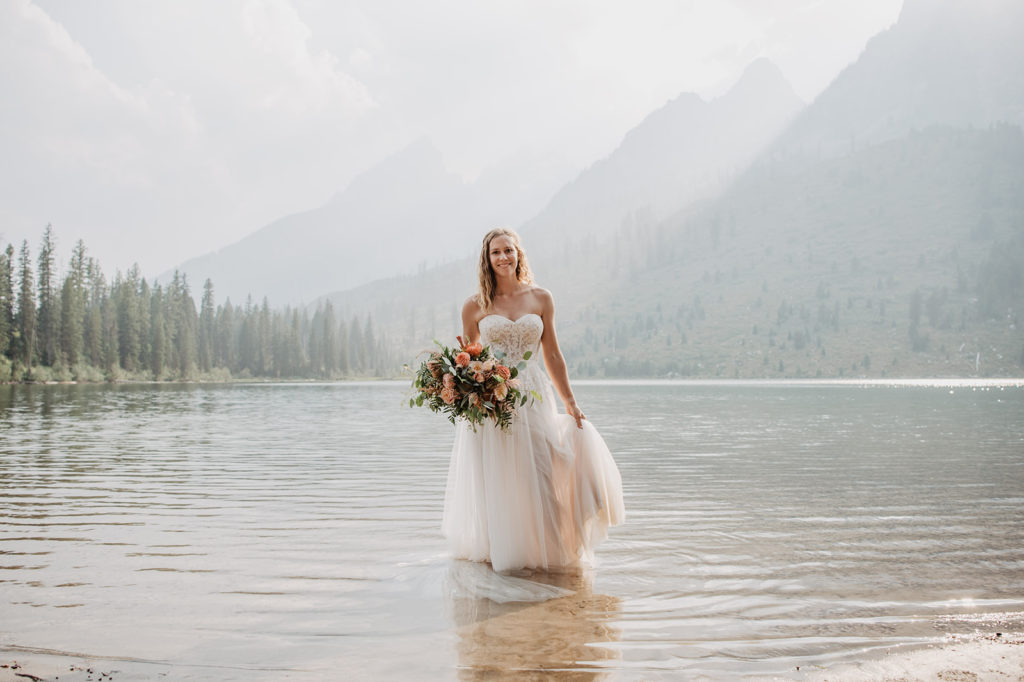 bride in the Tetons walking out of a lake in her strapless sweetheart neckline a-line lace and tulle wedding gown while holding her wedding bouquet with the Tetons in the background behind the lake with tall pine trees