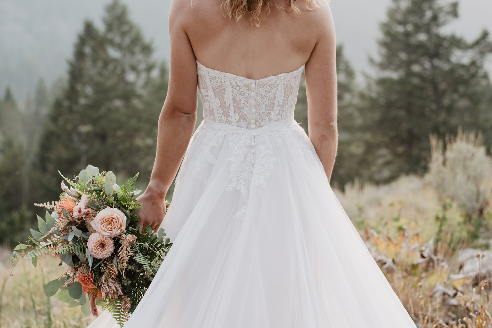 the back of a corset bodice wedding dress with the bride holding a bouquet of florals as she stands on a mountain top in Jackson Hole for her Jackson Hole wedding