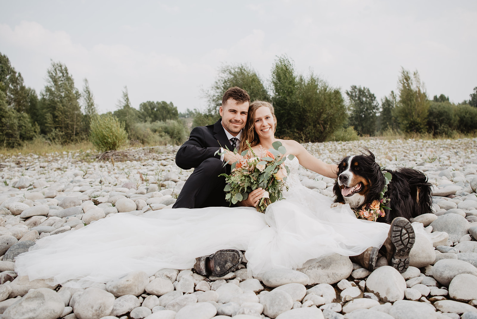 bride and groom sitting on rocks on the shore of a river in Jackson Hole with their dog