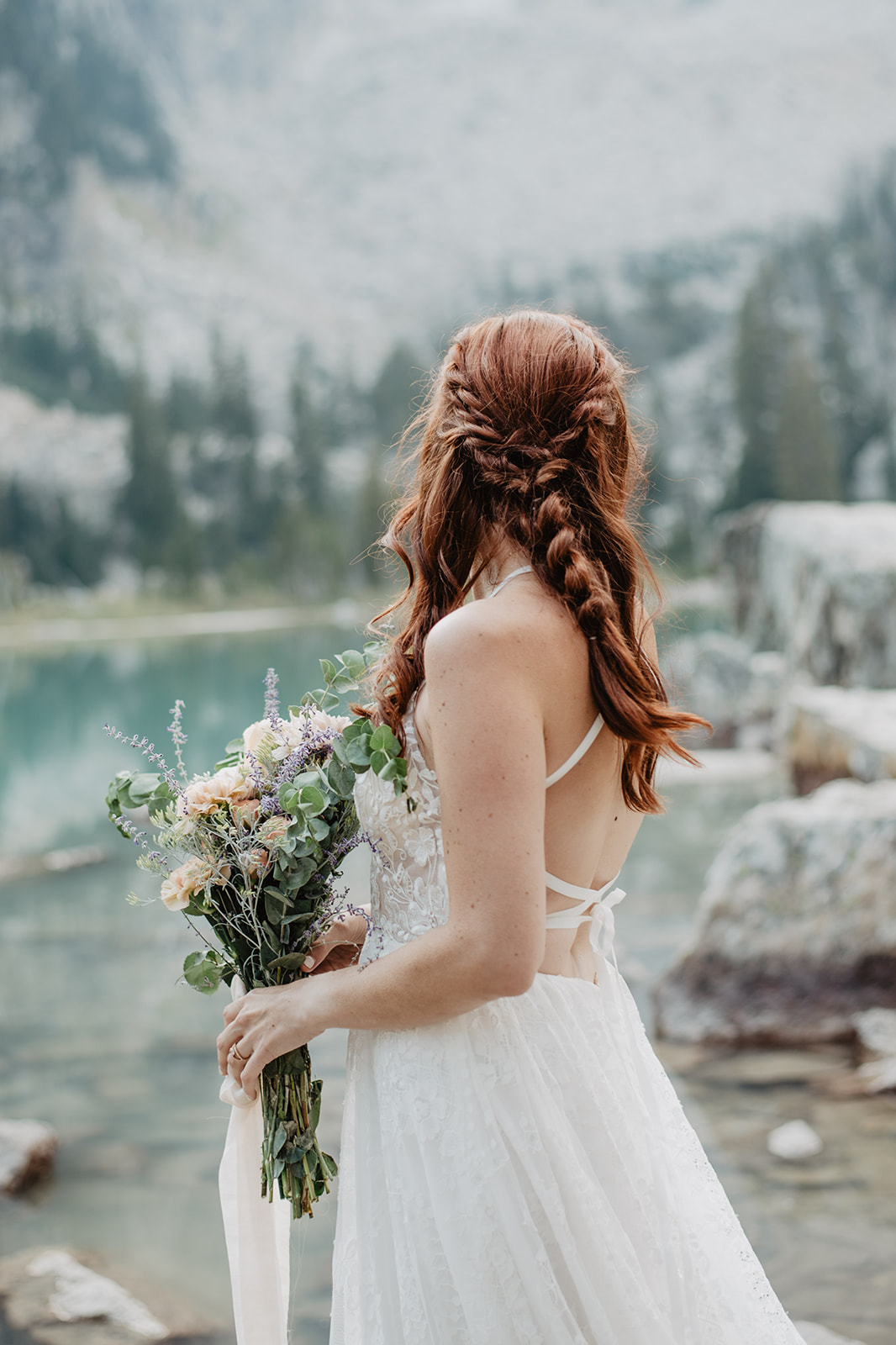 fall adventure elopement in the Tetons with the Tetons with the red headed bride holding a wild and free wedding bouquet and looking into an aqua blue glacier lake while wearing a lace gown