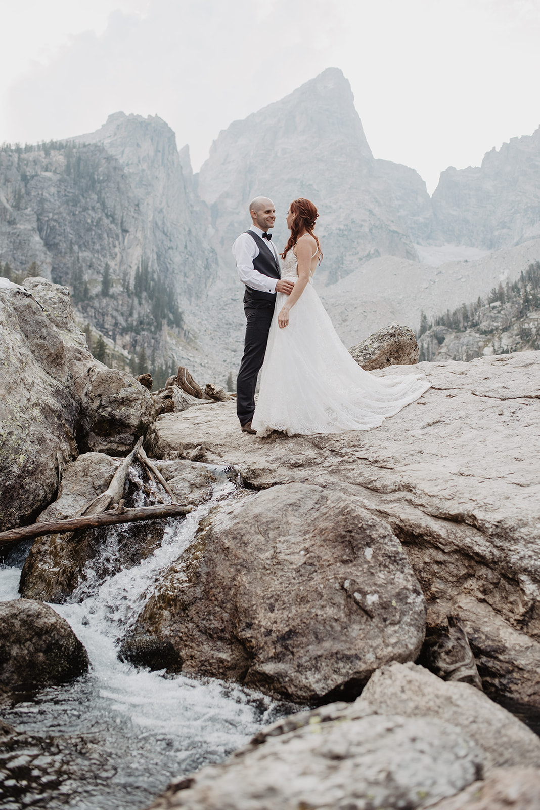 Teton adventure elopement in the Grand Tetons with a bride and groom standing on some rocks next to a lake with the Teton mountain range in the background