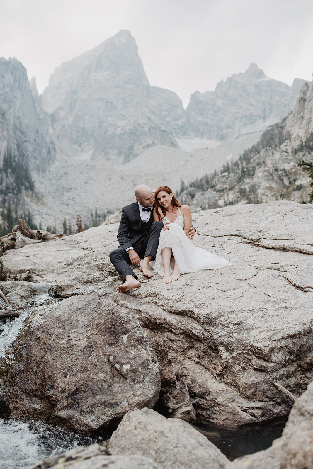 fall elopement in the Tetons with bride and groom sitting on the edge of a boulder cuddling each other as a small stream runs by them with the Teton mountain range behind them